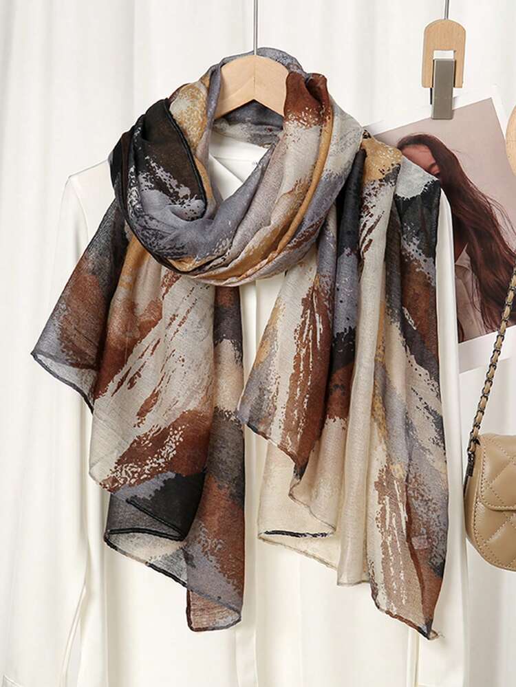 A lightweight scarf with an abstract painterly pattern in shades of brown, beige, gray, and black, draped over a white dress on a wooden hanger.