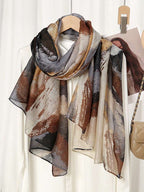 A lightweight scarf with an abstract painterly pattern in shades of brown, beige, gray, and black, draped over a white dress on a wooden hanger.