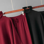 A close-up of knit garments hanging on a wooden hanger against a light gray wall. The most prominent item is a dark red turtleneck sweater, with a matching dark red skirt featuring a drawstring waist and a black turtleneck sweater partially visible behind it.