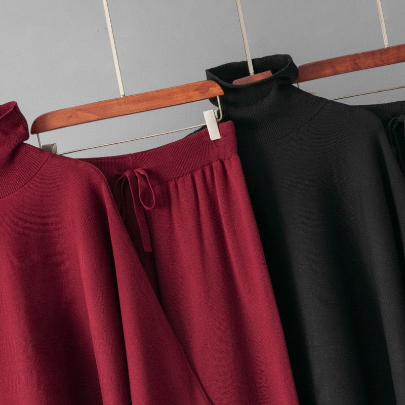 A close-up of knit garments hanging on a wooden hanger against a light gray wall. The most prominent item is a dark red turtleneck sweater, with a matching dark red skirt featuring a drawstring waist and a black turtleneck sweater partially visible behind it.