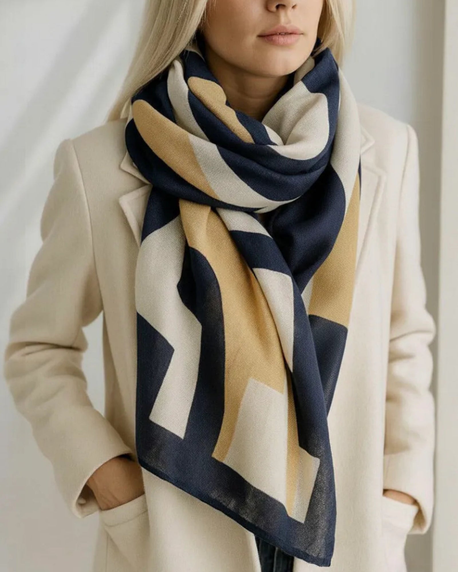 Person wearing a beige coat and a large scarf with geometric pattern on a plain background