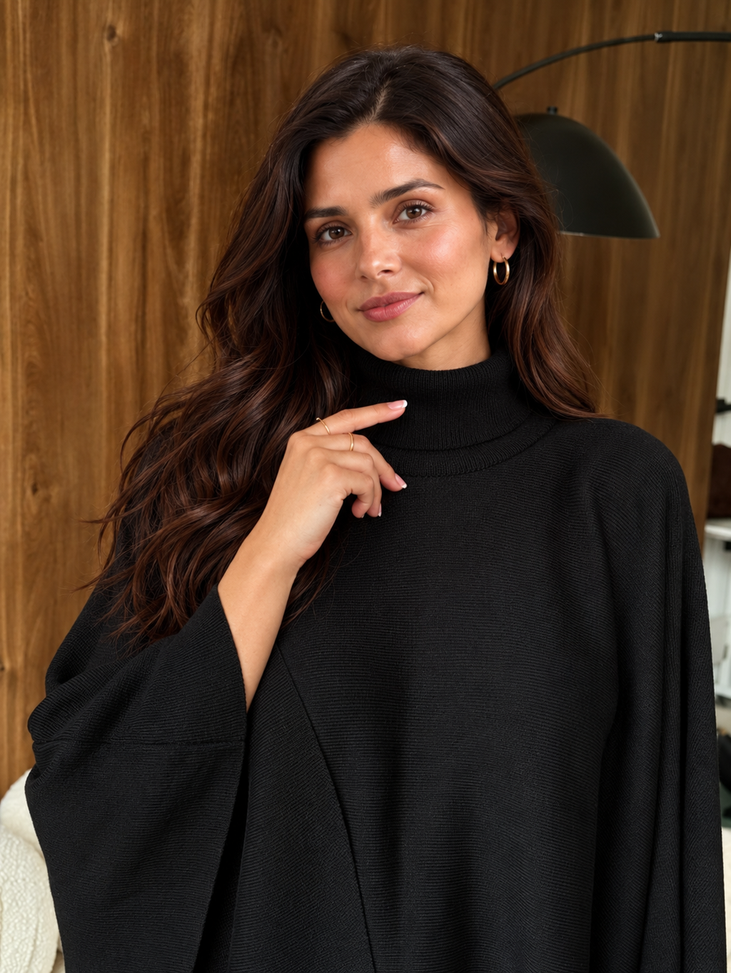 A woman with long dark brown hair wearing a black mock-neck knit top or poncho, posing in front of a wooden wall background.