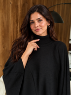 A woman with long dark brown hair wearing a black mock-neck knit top or poncho, posing in front of a wooden wall background.