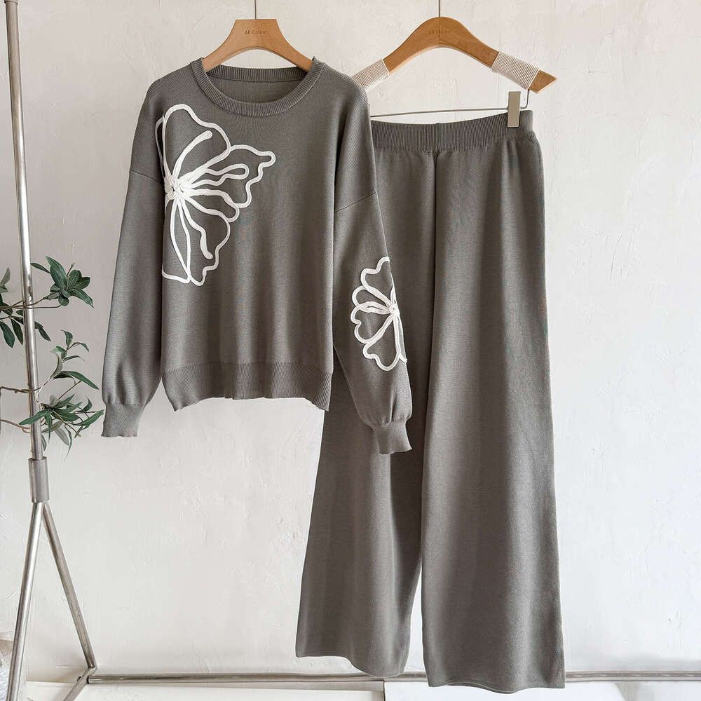 A two-piece women's lounge set in a muted gray color, featuring a crewneck sweatshirt with a large white floral outline on the front and matching wide-leg pants, both hanging on wooden hangers against a white wall.