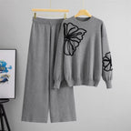 Gray sweater with butterfly design and matching pants on hangers against a gray background.