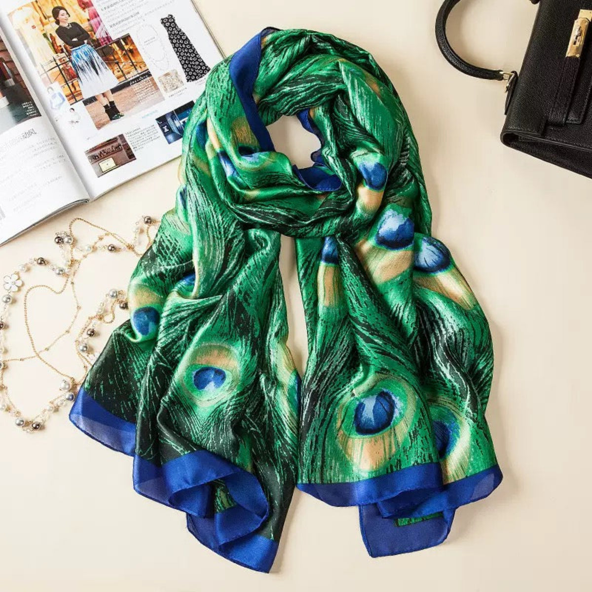 Green peacock feather patterned scarf on a light surface with a magazine and handbag in the background.