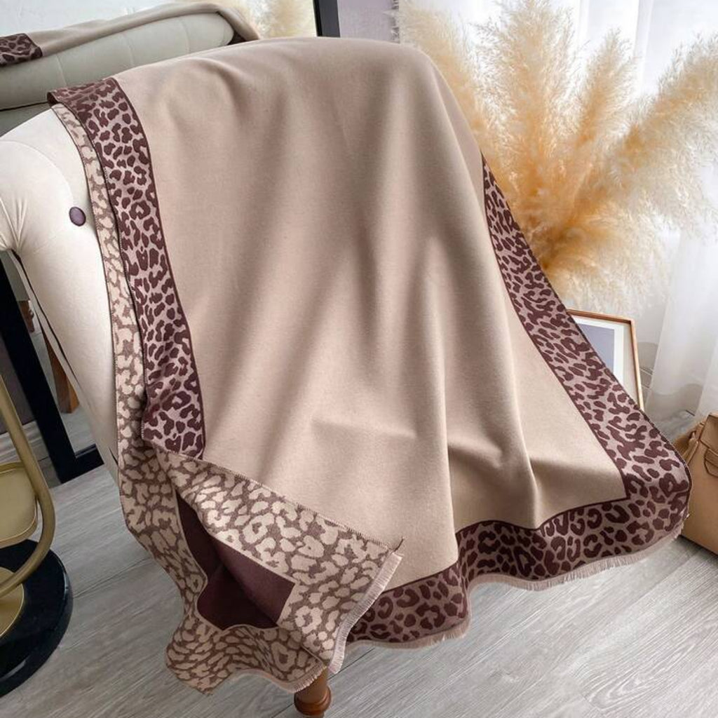 Beige blanket with leopard print border draped over a chair in a room with white curtains and pampas grass.