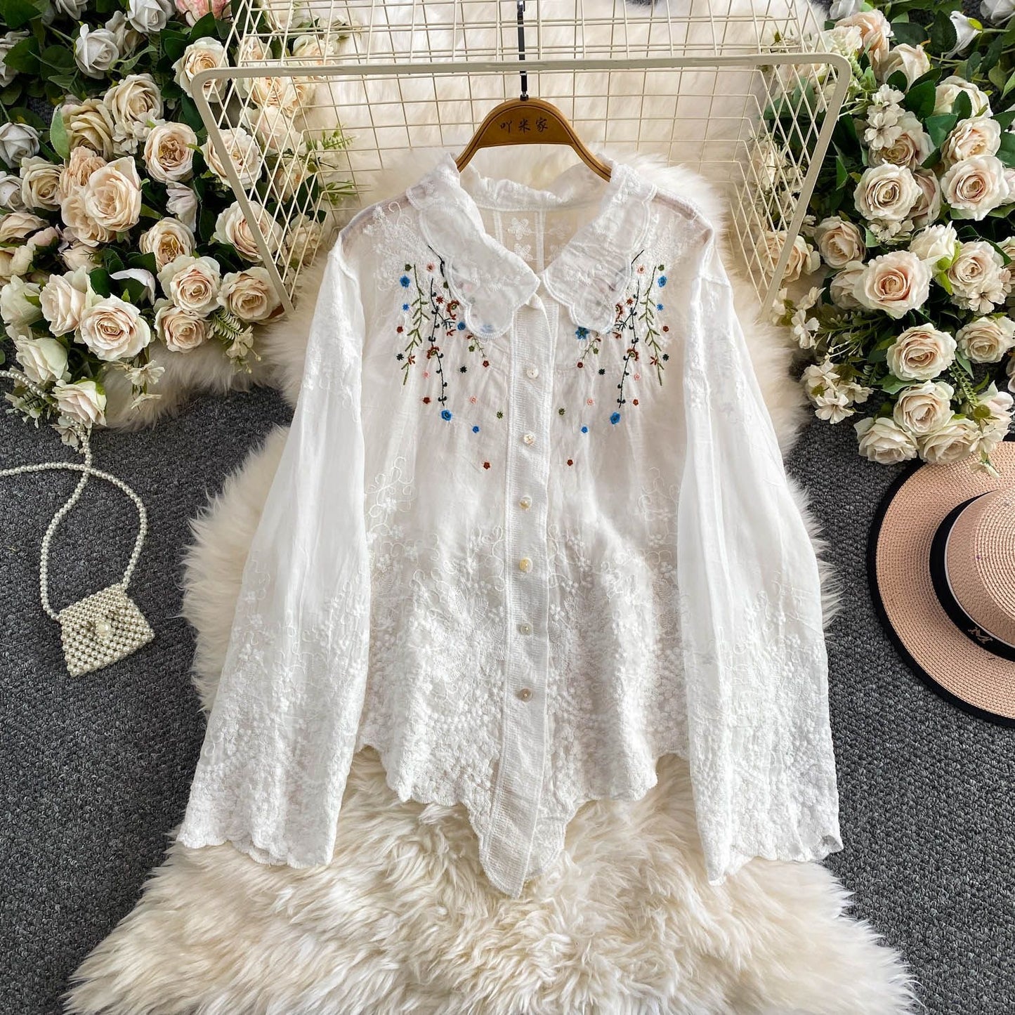 White embroidered blouse on a hanger with floral decorations and a hat in the background