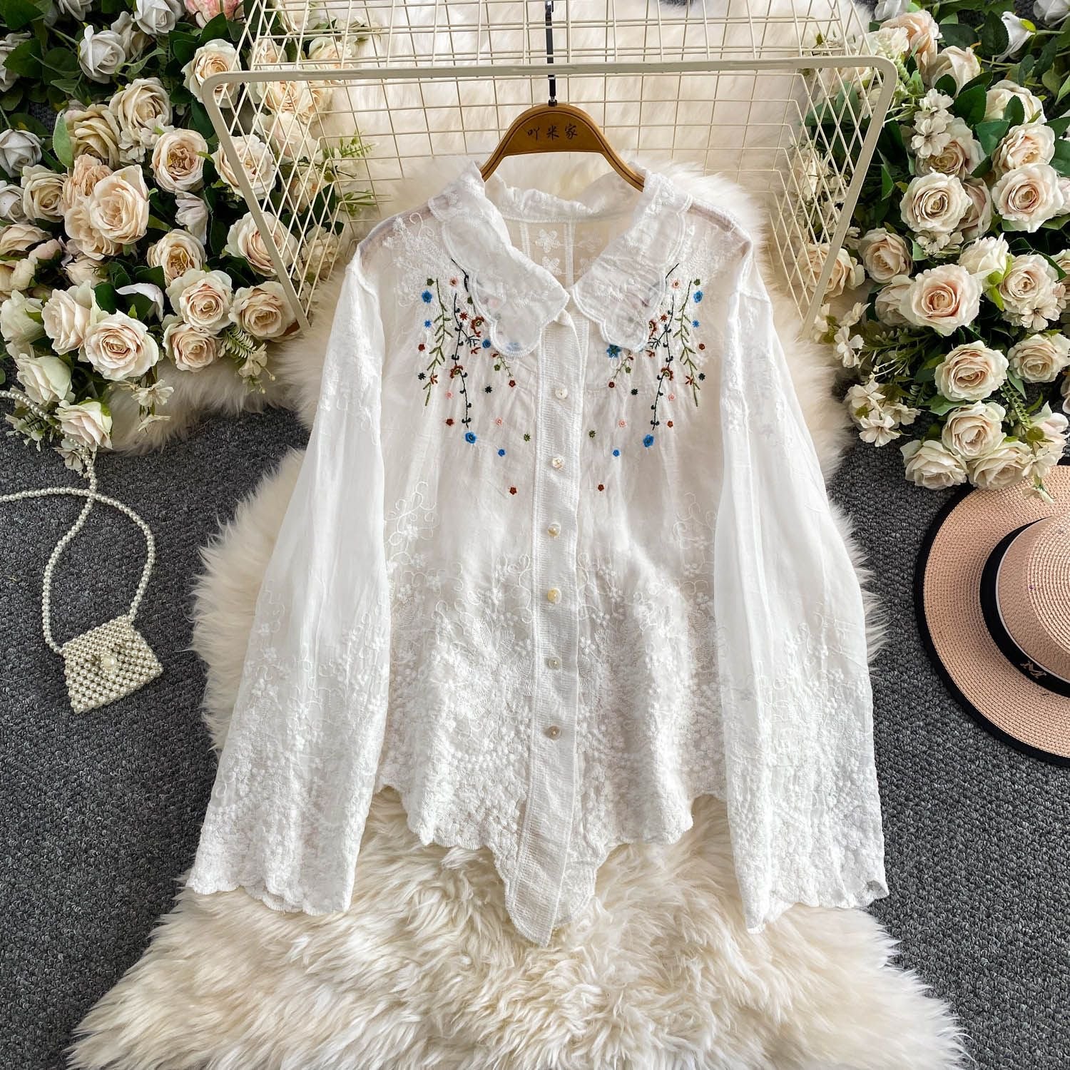 White embroidered blouse on a hanger with floral decorations and a hat in the background