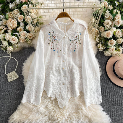 White embroidered blouse on a hanger with floral decorations and a hat in the background