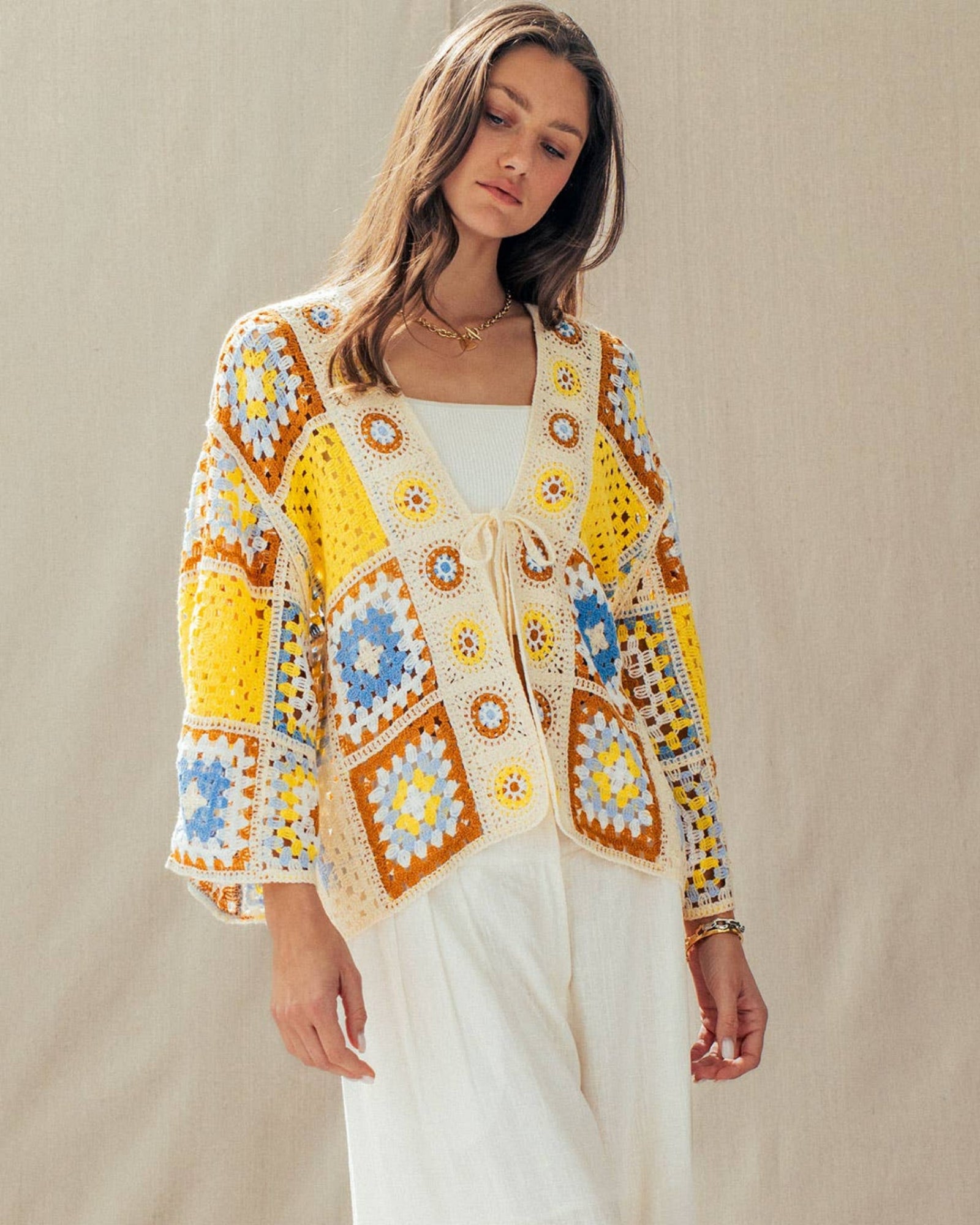 Woman wearing a colorful crochet cardigan and white pants against a beige background