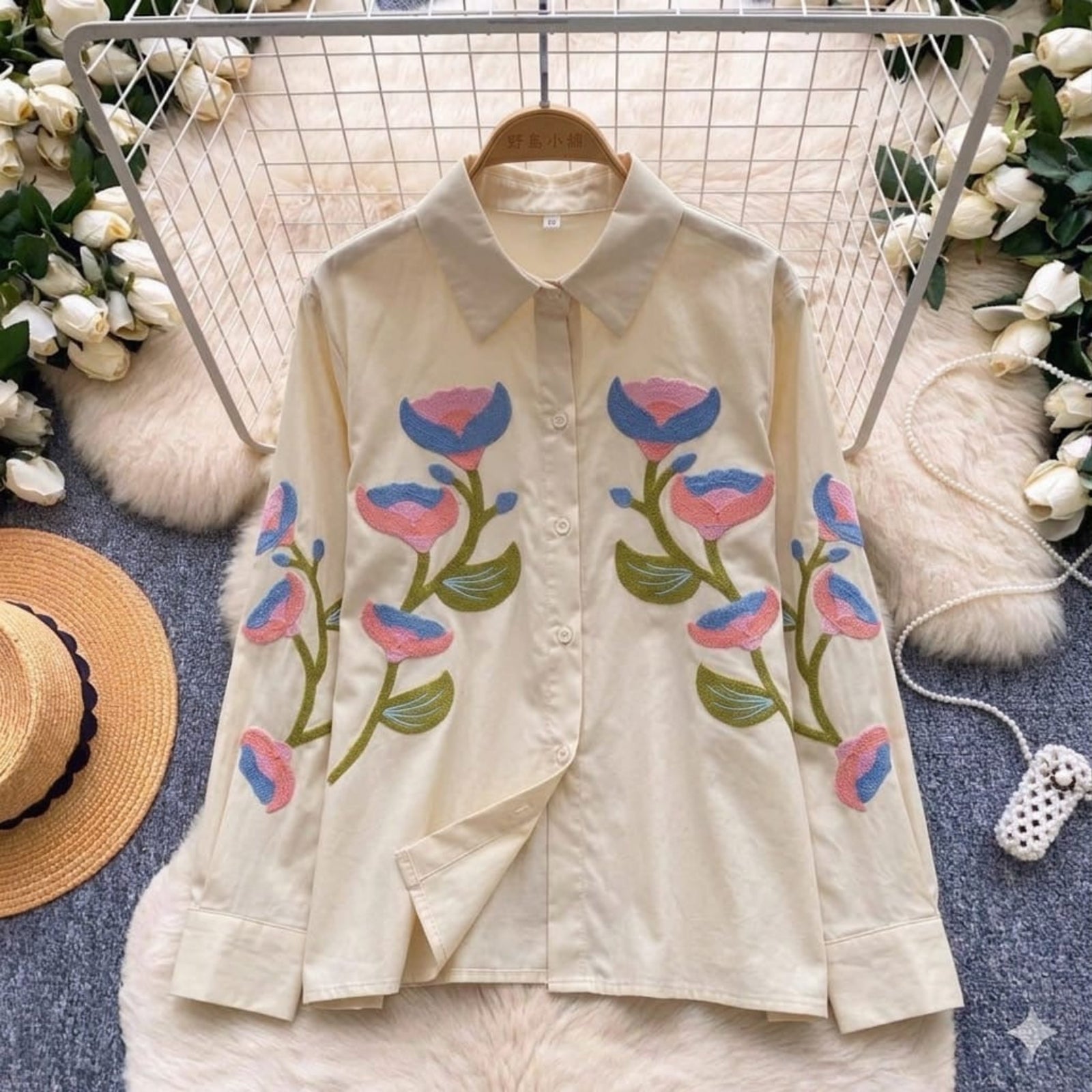 Beige shirt with floral embroidery on a hanger with flowers and a hat in the background