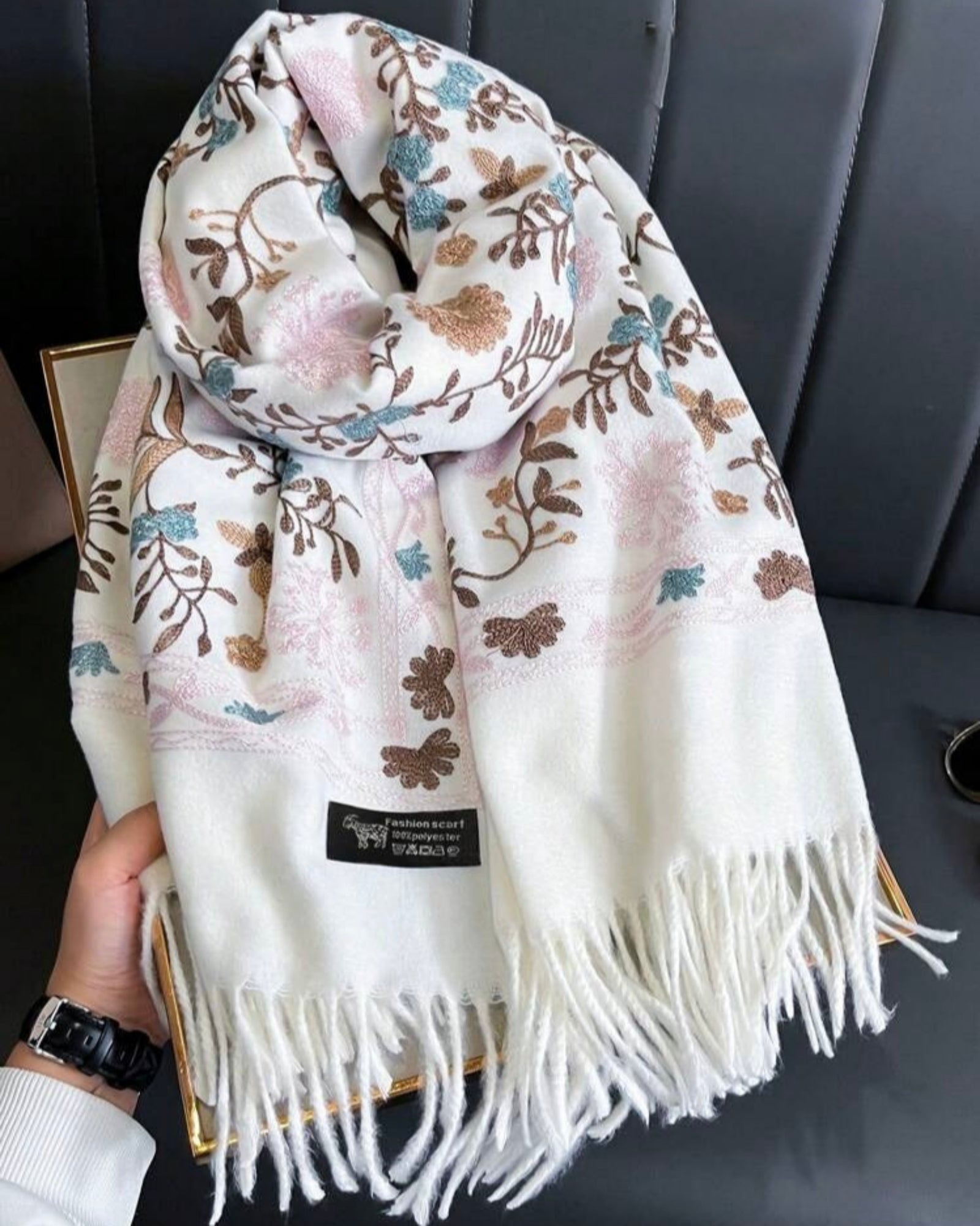 Floral patterned scarf with tassels held by a hand against a gray background