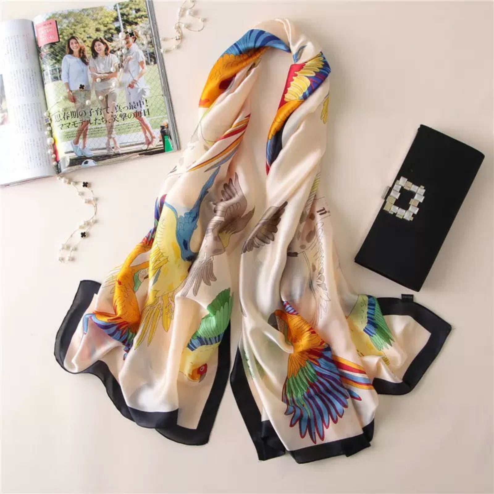 Colorful scarf with bird design on a white surface next to a magazine and black clutch.
