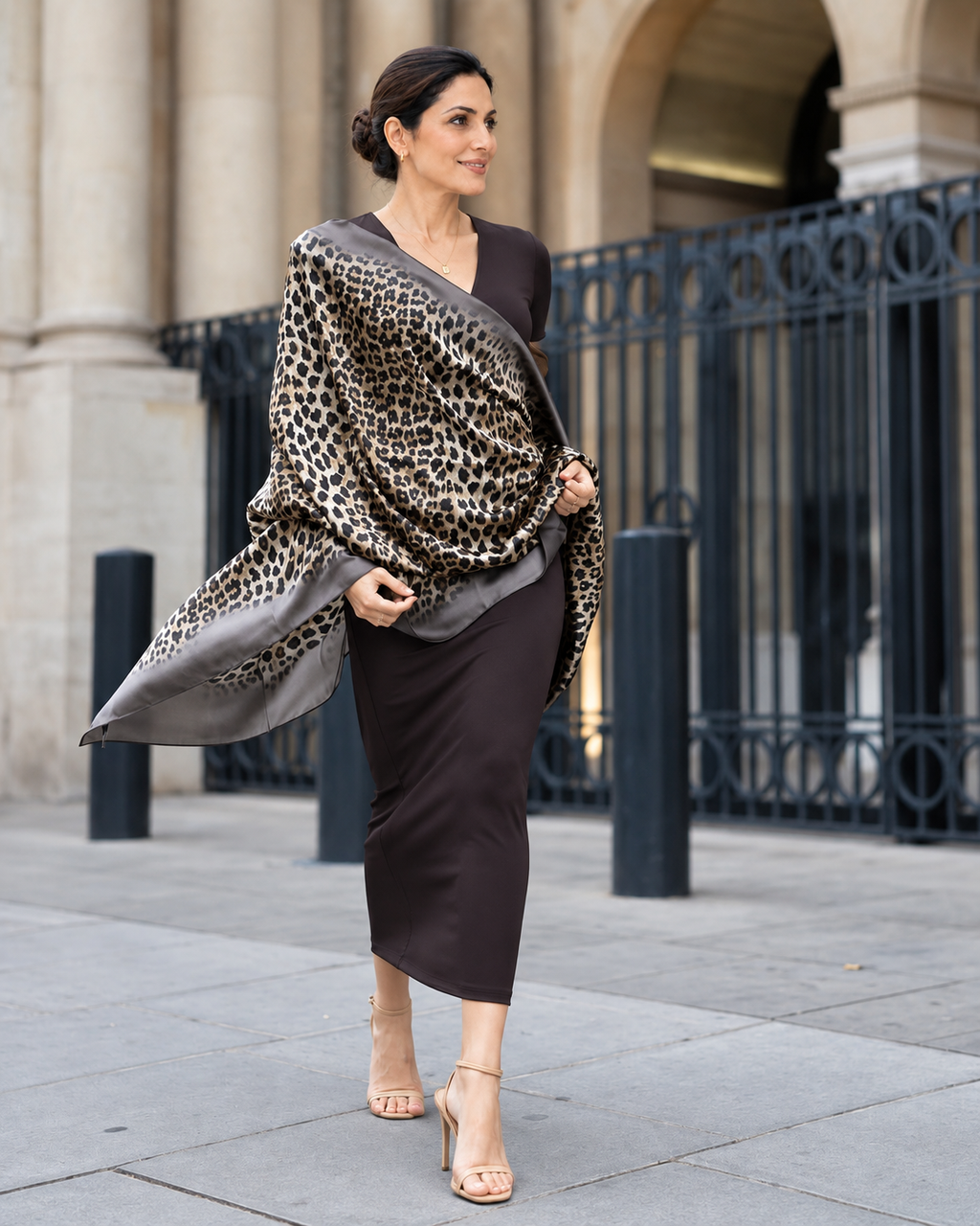 A woman stands outdoors in front of a stone building and black metal fence, wearing a dark brown midi dress and a leopard-print shawl draped over her shoulders.