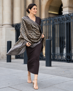A woman stands outdoors in front of a stone building and black metal fence, wearing a dark brown midi dress and a leopard-print shawl draped over her shoulders.