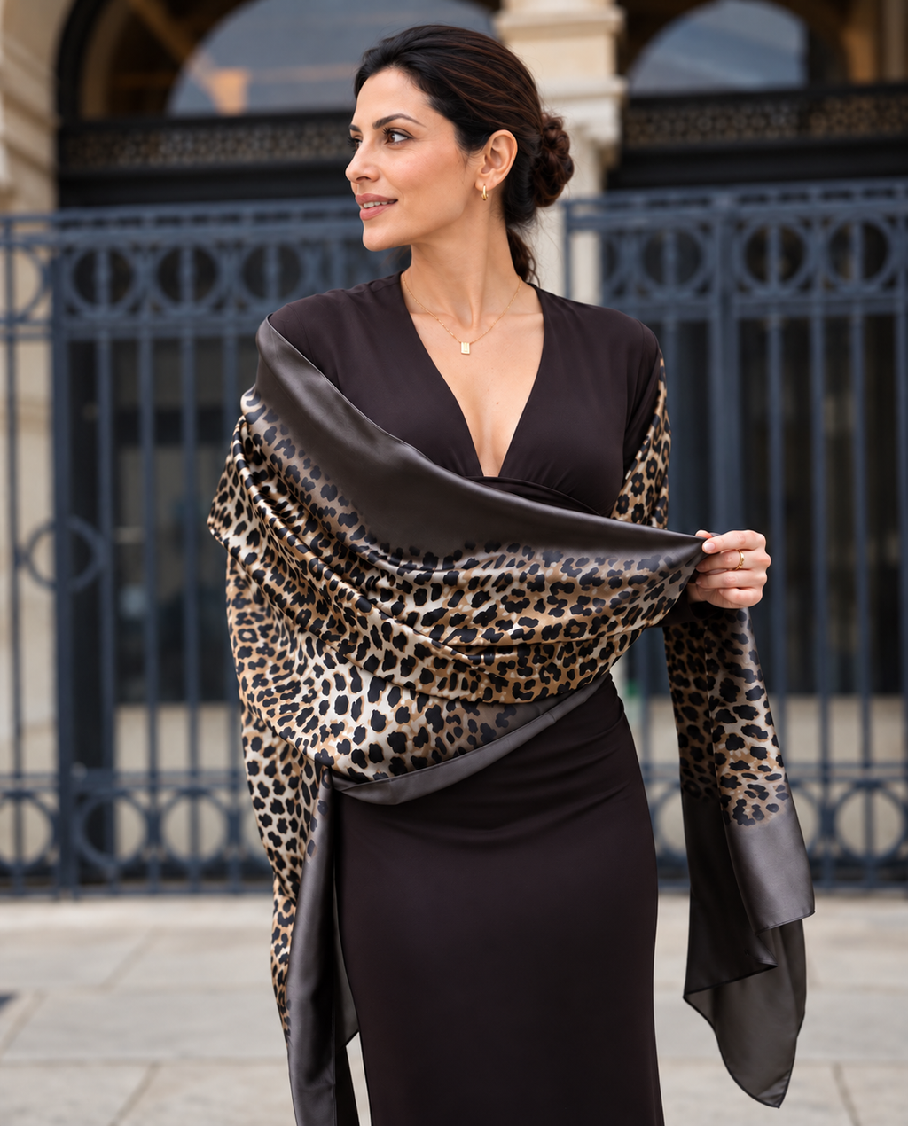 A woman wearing a dark brown dress and holding a leopard print silk scarf with dark brown satin borders, standing in front of a black wrought-iron gate.