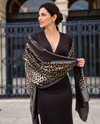 A woman wearing a dark brown dress and holding a leopard print silk scarf with dark brown satin borders, standing in front of a black wrought-iron gate.