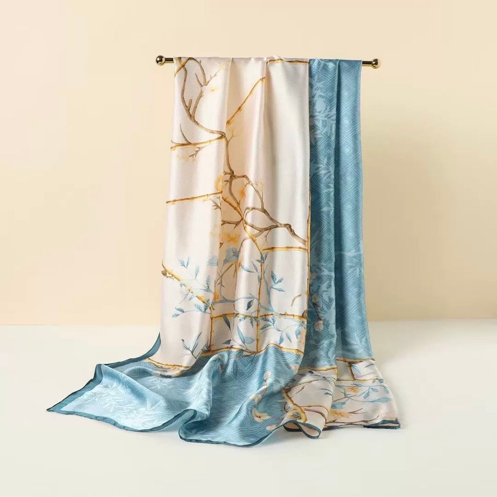 A long satin scarf draped over a gold rod, featuring a white and light blue floral pattern with gold branch details and a solid light blue border.