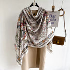 Westery Imperial Garden Bird & Bloom luxury silk scarf draped beautifully over a beige winter trench coat on a hanger, presenting a sophisticated, quiet luxury look for cold-weather accessorizing.
