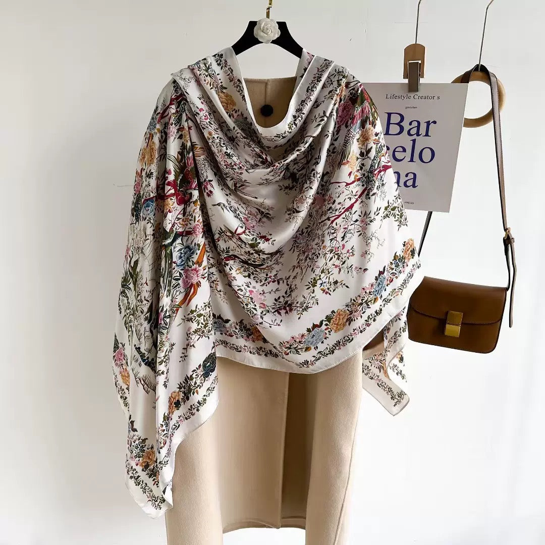 Westery Imperial Garden Bird & Bloom luxury silk scarf draped beautifully over a beige winter trench coat on a hanger, presenting a sophisticated, quiet luxury look for cold-weather accessorizing.
