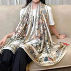 A model seated on a sofa wearing the Westery Imperial Garden Bird & Bloom premium silk scarf draped loosely over her shoulders, highlighting its generous size, elegant flow, and detailed floral border.
