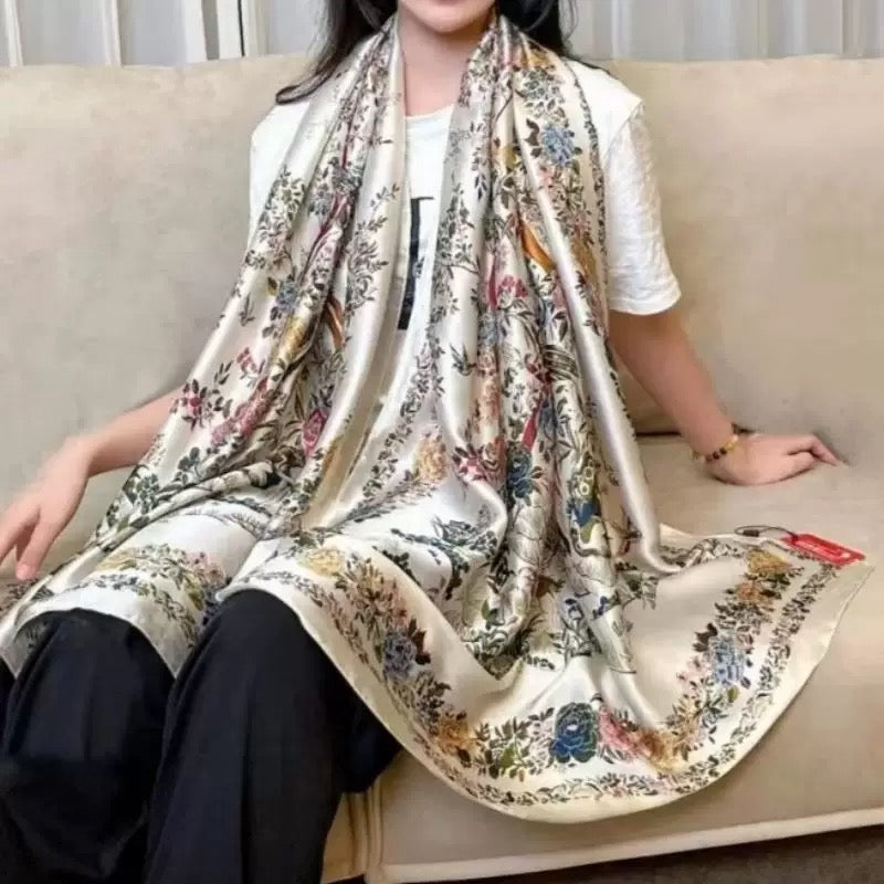 A model seated on a sofa wearing the Westery Imperial Garden Bird & Bloom premium silk scarf draped loosely over her shoulders, highlighting its generous size, elegant flow, and detailed floral border.
