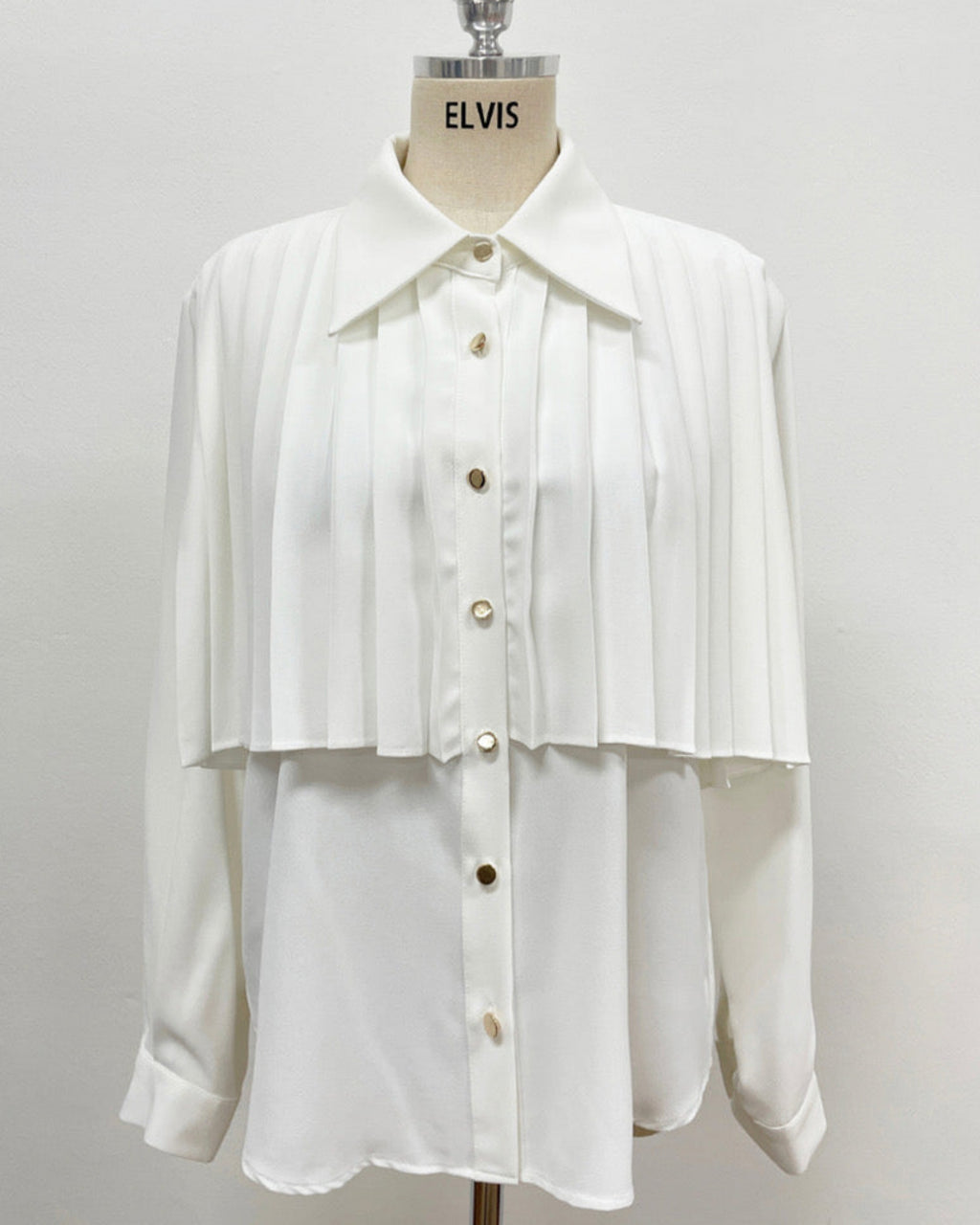 Detail view of white pleated cape collar shirt showing gold button closure and pleated overlay design